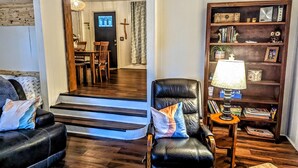 Uma sala de estar com um sofá de couro, uma cadeira de madeira, uma estante e uma escada que leva a outro cômodo.