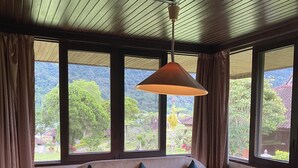 Una habitación con sofá y mesa, ventanas amplias con cortinas, y vista a zonas verdes.