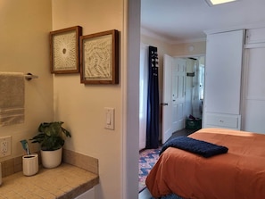 A hotel room with a bed, a bathroom with a towel rack, a potted plant, and framed artwork on the wall.