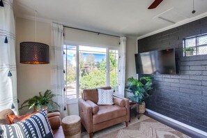 En moderne stue med sofa, sofabord, fjernsyn monteret på en murstensvæg og et stort vindue med udsigt til grønne omgivelser.