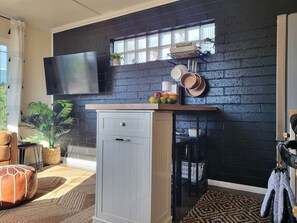 A modern kitchen with a black brick wall, a flat-screen TV mounted above the counter, and a window with a view. - Chic, Playful Boho Bungalow Near Everything! (La Mesa)