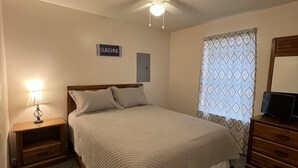 A bedroom with a bed, a nightstand with a lamp, a ceiling fan, a window with patterned curtains, and a wall-mounted TV. - STEPS FROM SAND (Gulf Shores)