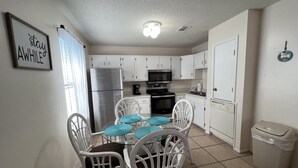 A compact kitchen with white cabinets, a stainless steel refrigerator, a microwave, and a stove. There is a dining table with four white chairs and blue cushions. A trash bin is visible to the right. - STEPS FROM SAND (Gulf Shores)
