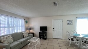A living room with a sofa, a coffee table, a TV, a dining table with chairs, and a door. - STEPS FROM SAND (Gulf Shores)