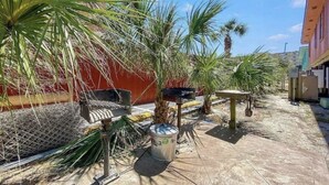 A courtyard with palm trees, a metal trash bin, and a red building in the background. - STEPS FROM SAND (Gulf Shores)