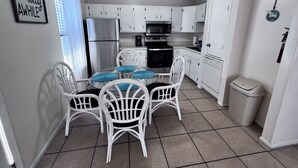 A compact kitchen with white cabinets, a stainless steel refrigerator, a microwave, and a stove. There is a dining table with white chairs and blue cushions. A trash bin is visible in the corner. - Convenient to everything! Only steps to the beach. (Gulf Shores)