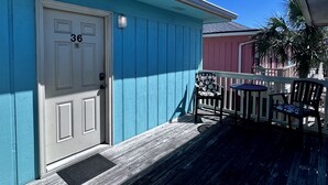 A blue building with a white door marked '36', a small porch with a chair and a table, and a palm tree in the background. - Convenient to everything! Only steps to the beach. (Gulf Shores)
