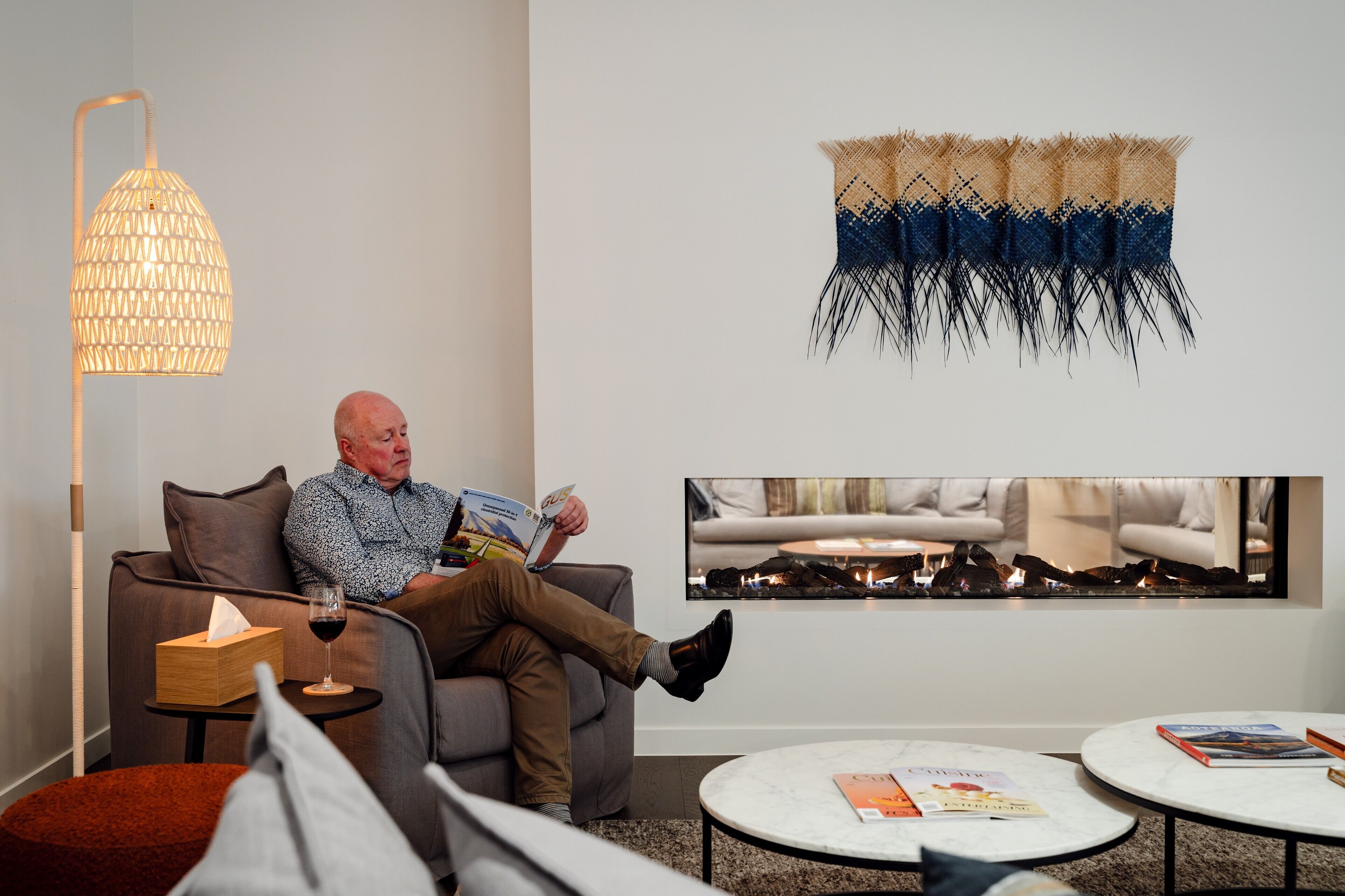 Un hombre leyendo un libro en un sofá en una sala de estar moderna con chimenea, una obra de arte en la pared y una mesa de centro redonda con libros.