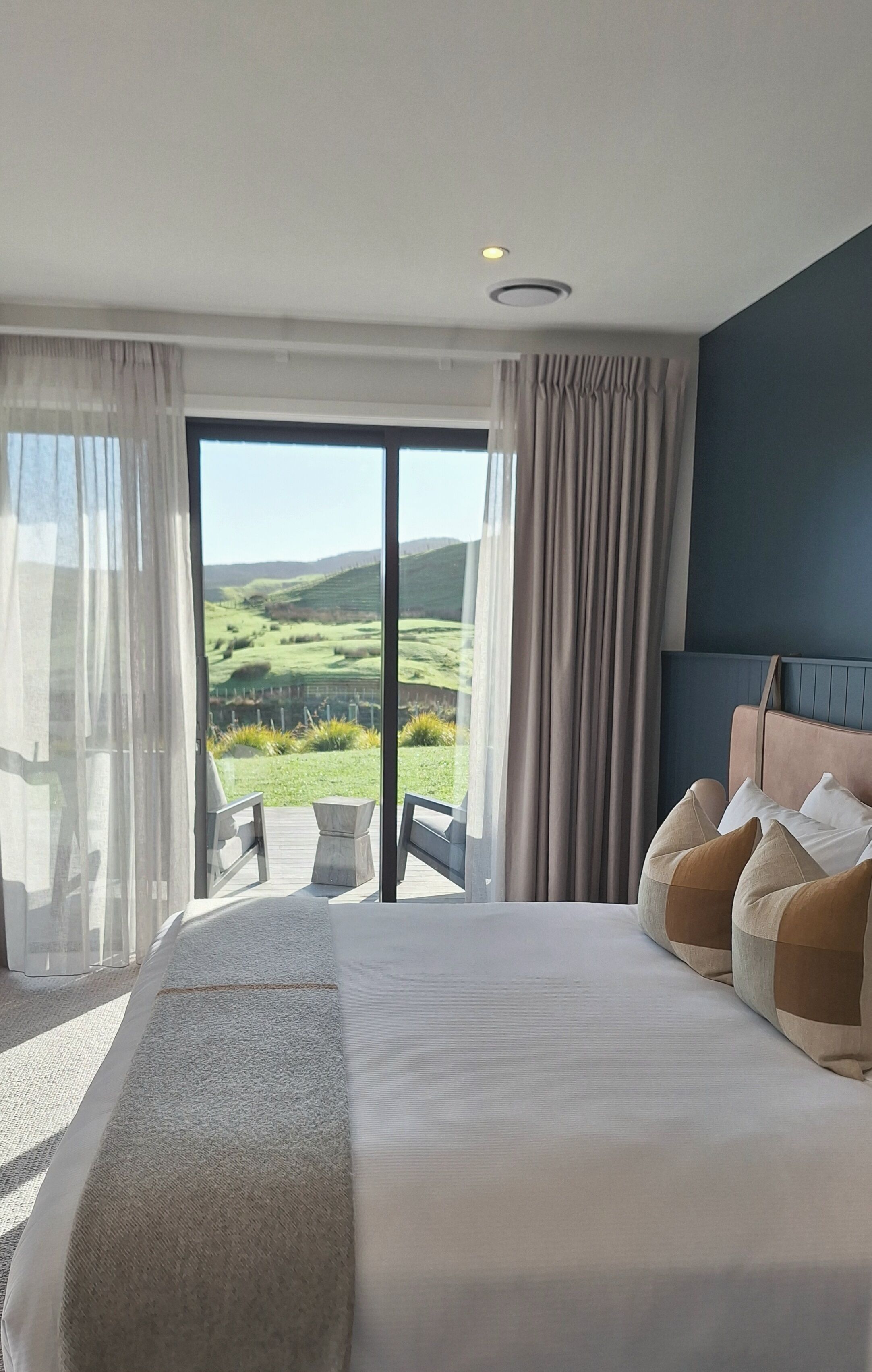 A hotel room with a large bed, a view of a green landscape through a sliding door, and a balcony with a table and chairs.