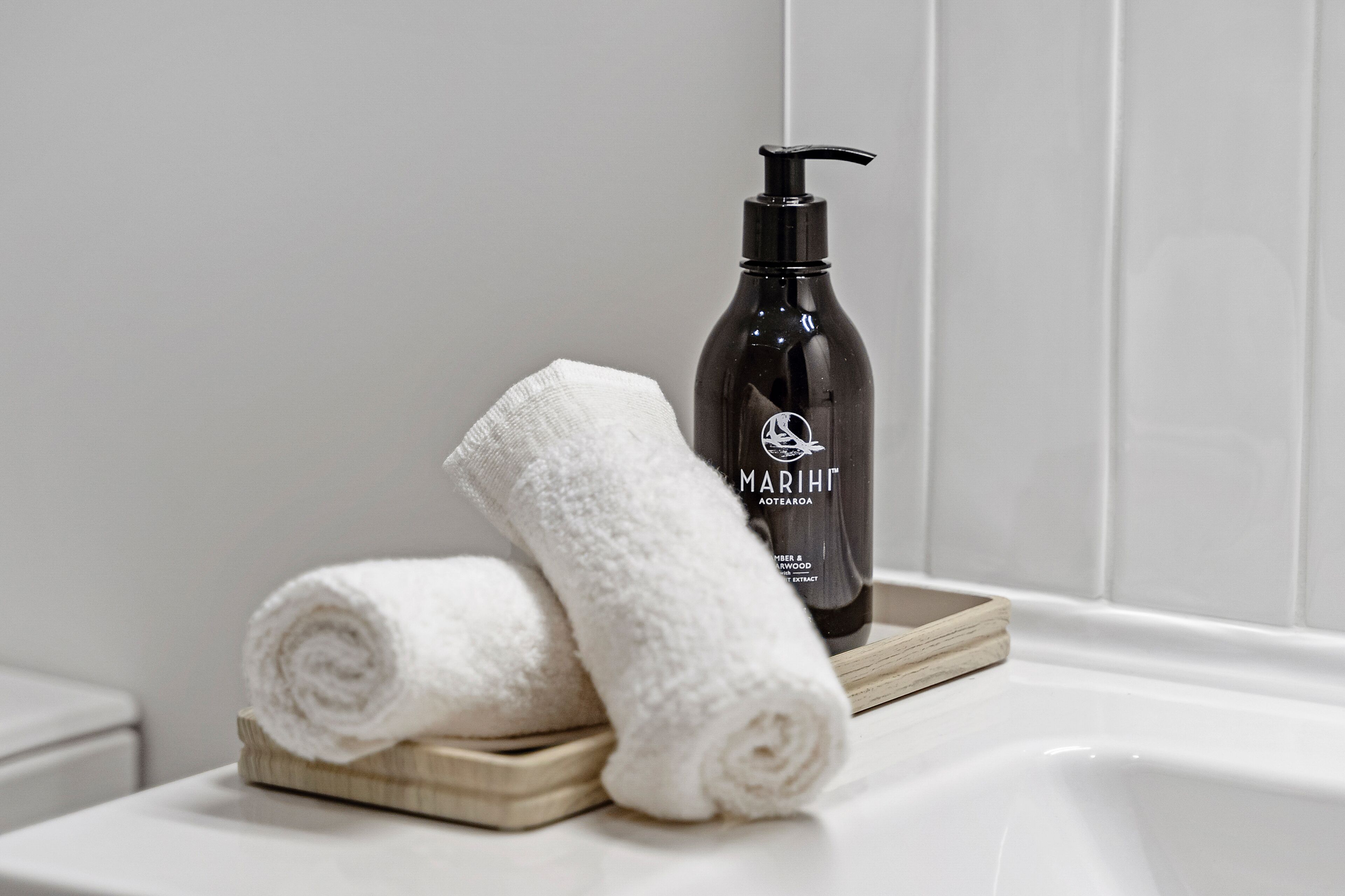 A bathroom with a white bathtub, a black bottle of Marijke Aoteaqua body wash, and two white towels on a wooden tray.