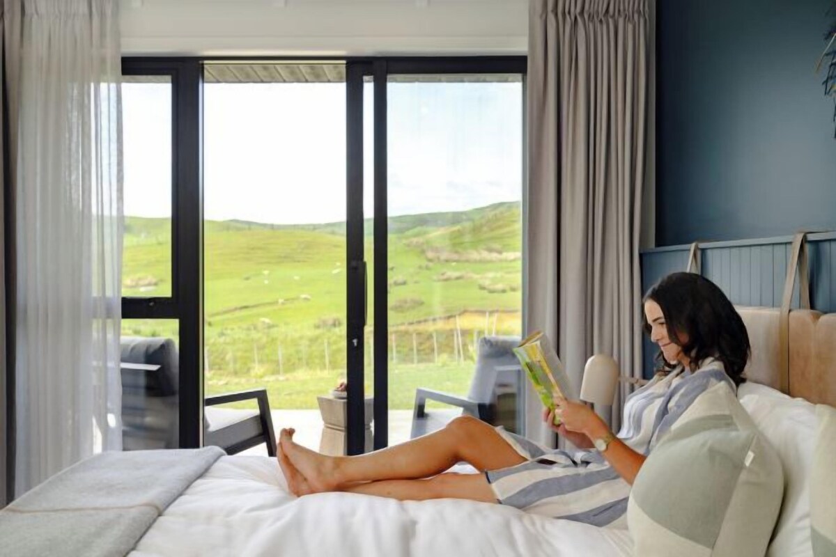 A room with a large window, a bed, and a person reading a book.