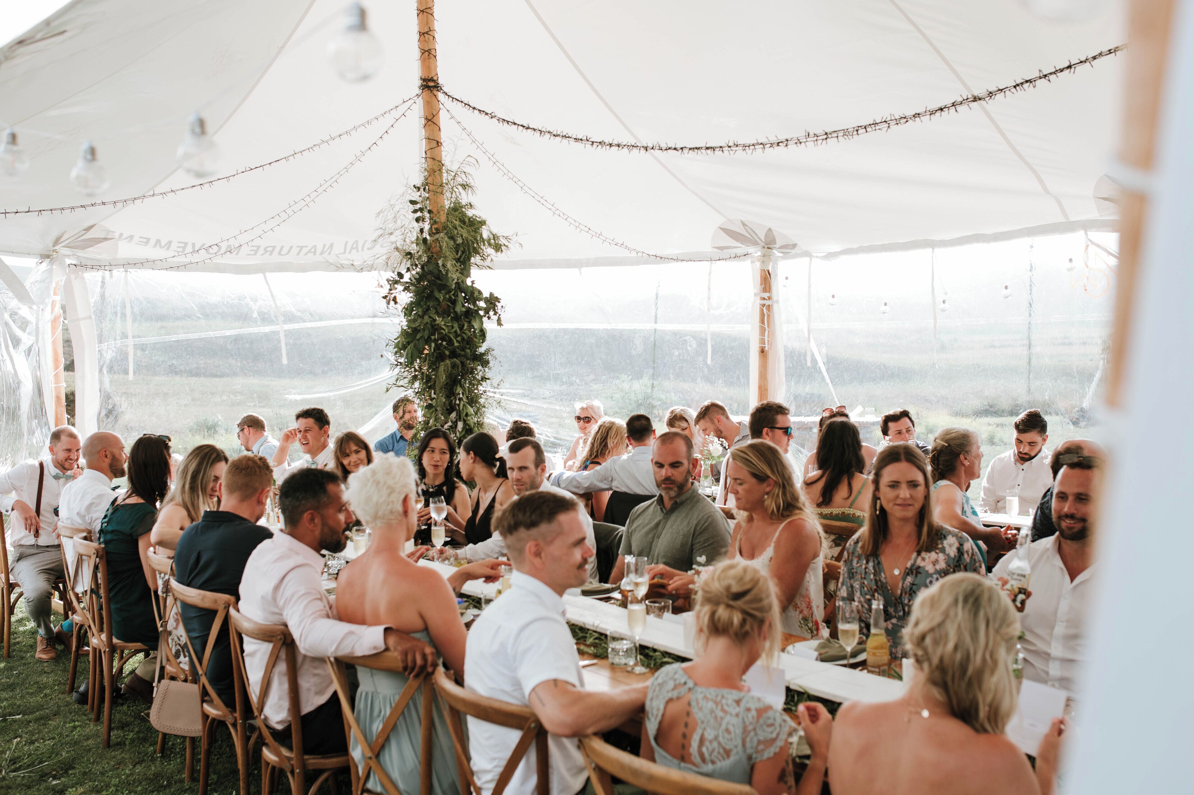 A tented outdoor dining area with tables and chairs, surrounded by greenery and a view of the landscape.