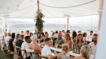 A tented outdoor dining area with tables and chairs, surrounded by greenery and a view of the landscape.