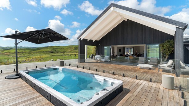 A modern outdoor pool area with a hot tub, wooden deck, and a covered seating area with glass railings. - Te Karaka Lodge (Waikaretu)