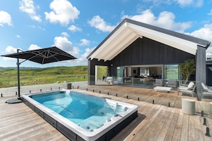 Un área de piscina moderna al aire libre con hidromasaje, terraza de madera y zona de estar cubierta con barandillas de vidrio.