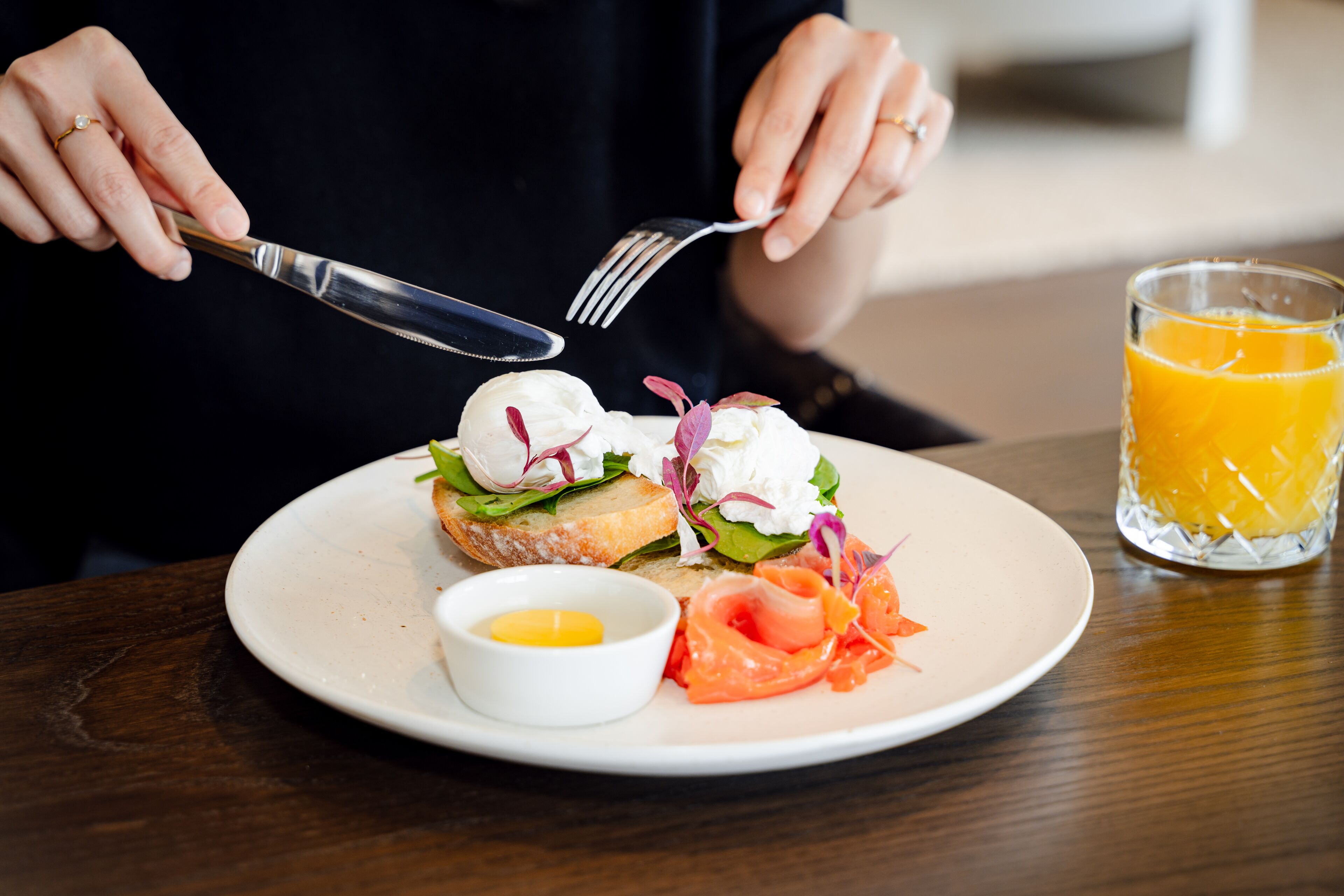 A person holding a fork and knife over a plate with a sandwich, a small bowl of sauce, and a glass of orange juice.