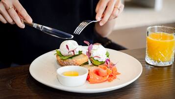 A person holding a fork and knife over a plate with a sandwich, a small bowl of sauce, and a glass of orange juice.