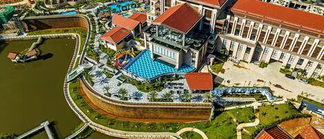 An aerial view of a hotel with a swimming pool, a large building with a red roof, and a surrounding landscaped area.