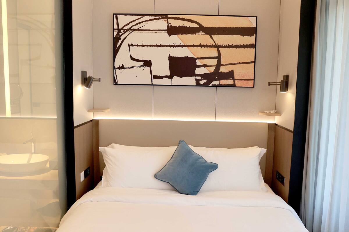 A hotel room with a bed, a wall-mounted abstract painting, a bedside table, and a nightstand.