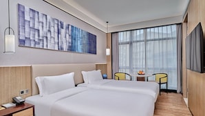 Habitación de hotel con dos camas, un ventanal con cortinas translúcidas, un televisor de pantalla plana, una mesita con frutero, y una obra de arte colgada en la pared.