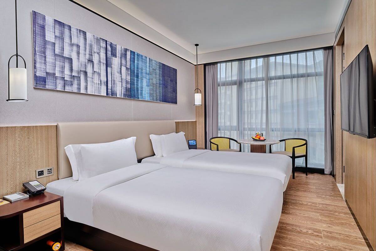 A hotel room with two beds, a large window with sheer curtains, a flat-screen TV, a small table with a fruit bowl, and a wall-mounted artwork.