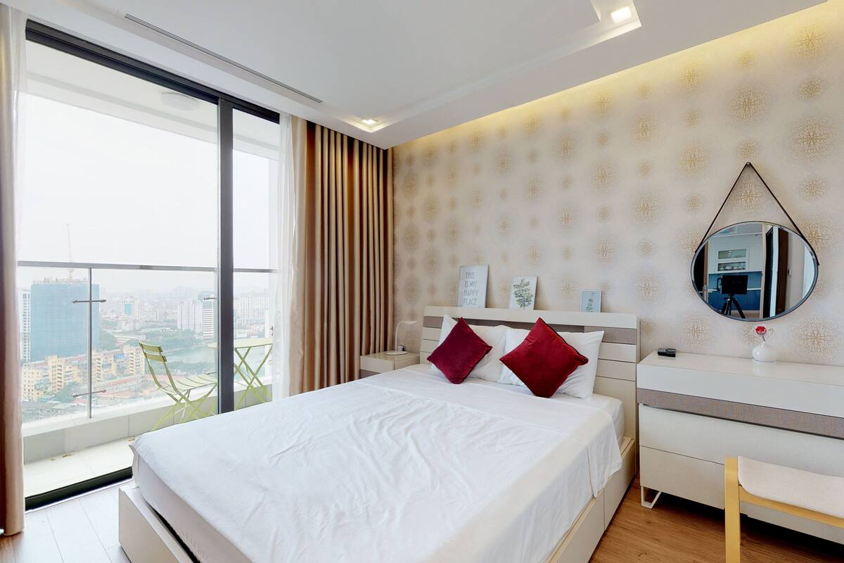 Een moderne hotelkamer met een groot bed, nachtkastje, spiegel en uitzicht op de stad door een groot raam.