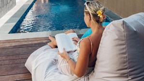 Una persona leyendo un libro junto a una piscina, con una casa moderna al fondo.