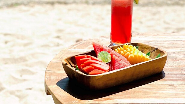Una mesa en la playa con una bebida y un cuenco de fruta.
