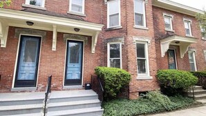 A two-story brick building with a front porch, a black front door, and a small garden area with trimmed bushes.