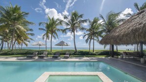 A pool area with lounge chairs and umbrellas, surrounded by palm trees and a view of the ocean. - Ocean Front Luxury Villa in Golf and Beach Resort (Punta Cana)