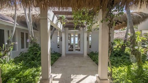 A covered walkway with columns and a thatched roof, surrounded by lush greenery and leading to a building with large windows. - Ocean Front Luxury Villa in Golf and Beach Resort (Punta Cana)