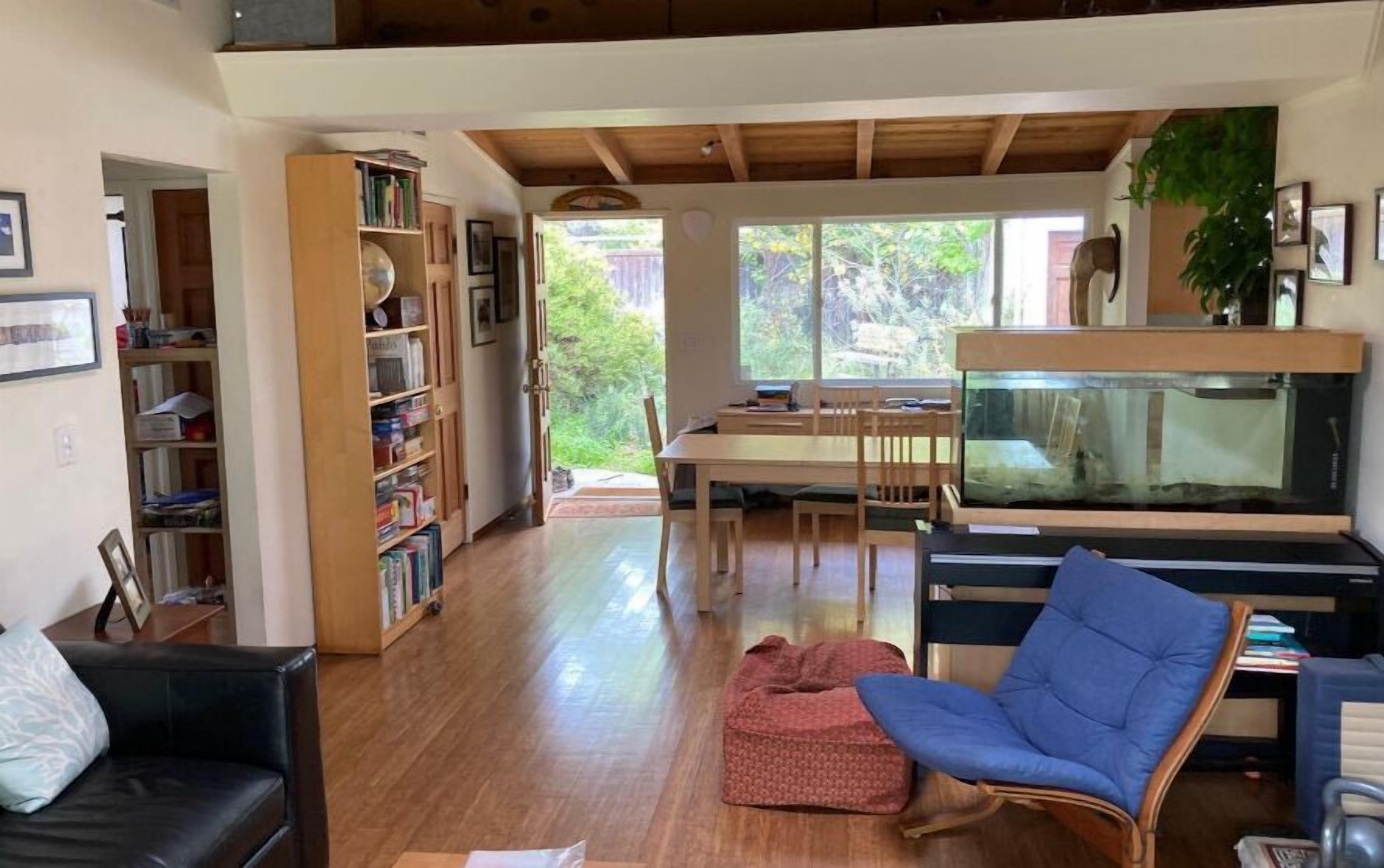 Amplia sala de estar con piso de madera, una mesa de comedor con sillas, una estantería, un sillón azul y un acuario.