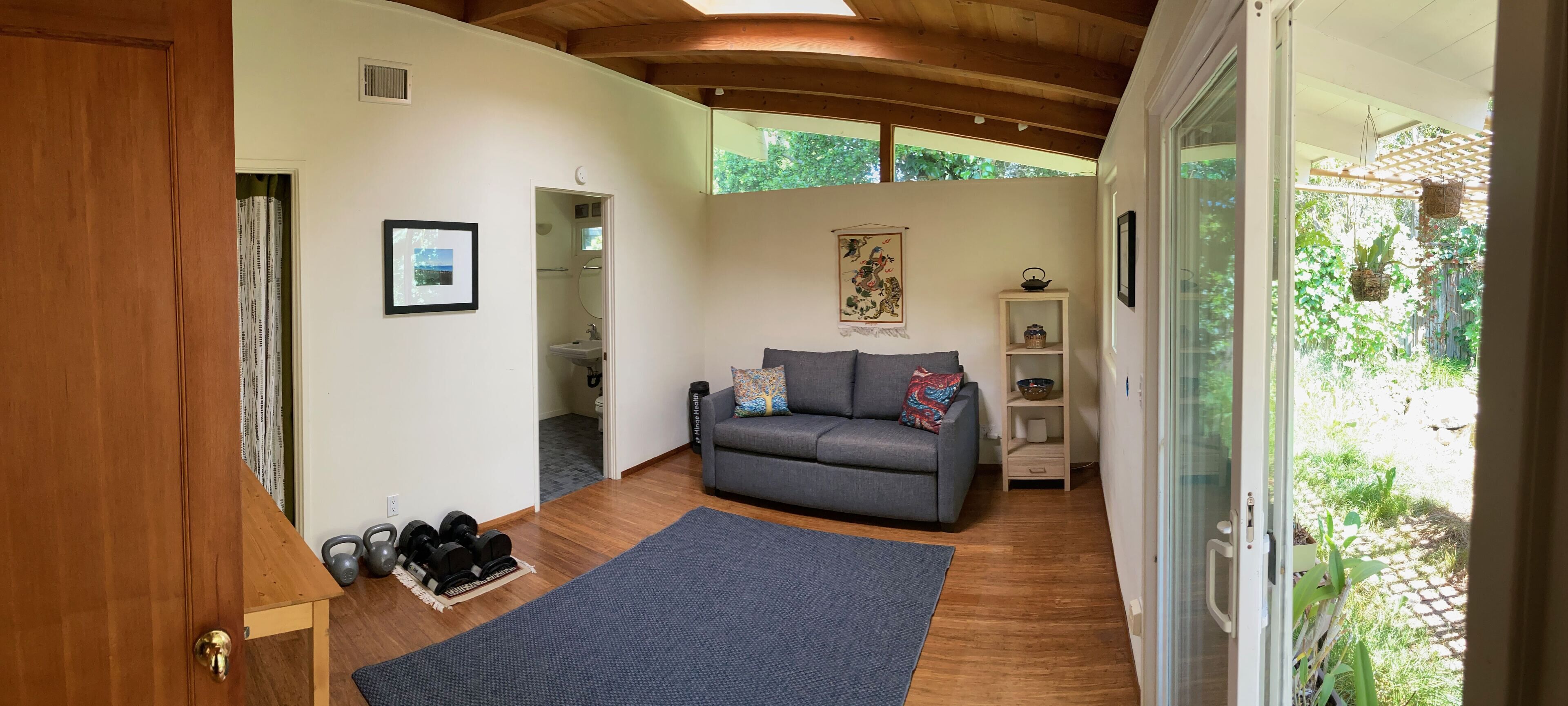 Ein gemütliches Wohnzimmer mit grauem Sofa, einer kleinen Ablage mit Dekoration, einem gerahmten Bild an der Wand und Blick ins Grüne.