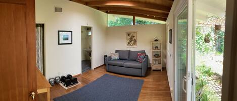 Ein gemütliches Wohnzimmer mit grauem Sofa, einer kleinen Ablage mit Dekoration, einem gerahmten Bild an der Wand und Blick ins Grüne.