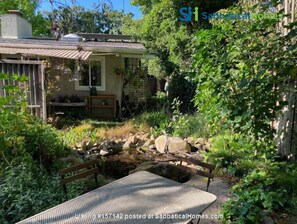 Escape to a serene garden oasis with a rustic bench and tranquil pond, surrounded by lush greenery and a charming country-style home.