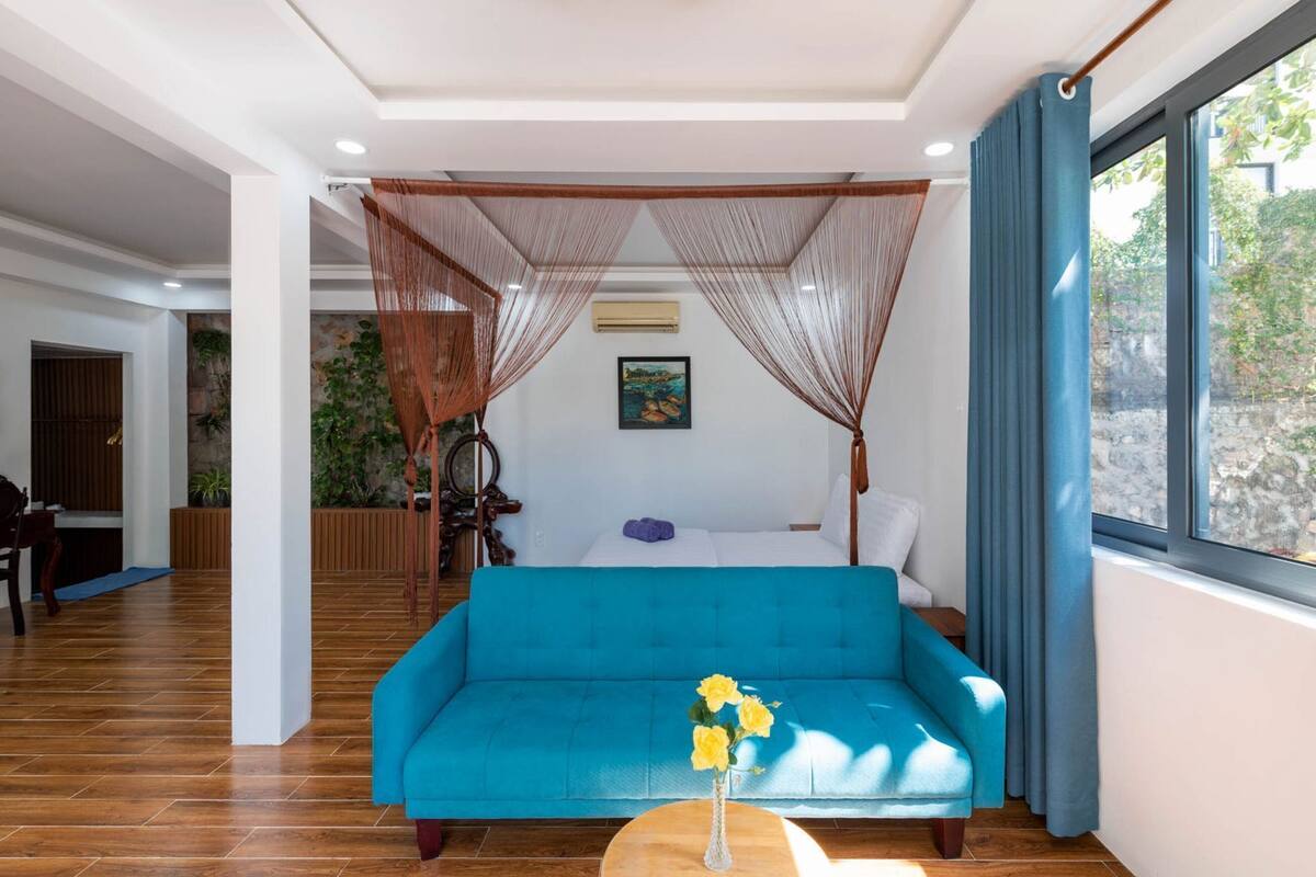 A modern room with a blue sofa, a canopy bed, a dining area, and a view of greenery outside.