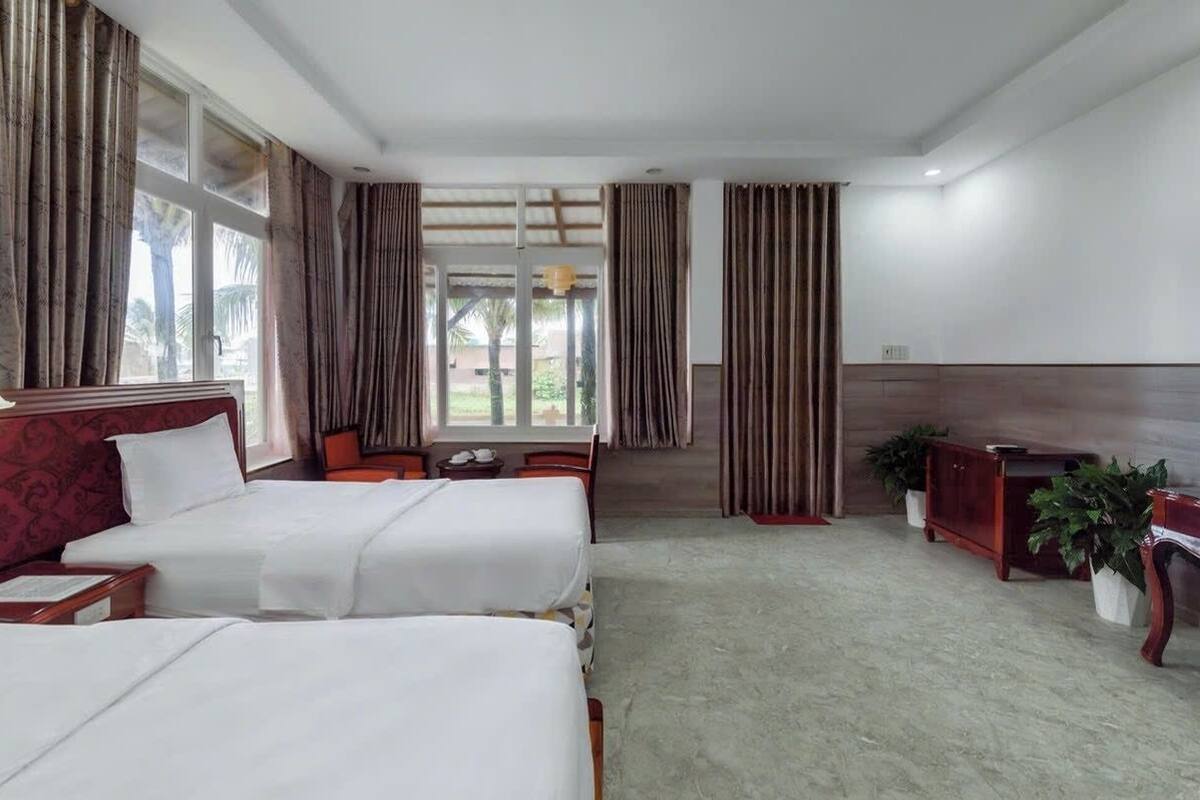 A hotel room with two beds, a desk, a chair, a small table, and a potted plant.
