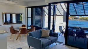 A modern living room with a grey sofa, a dining table with chairs, and large windows offering a view of the outside.