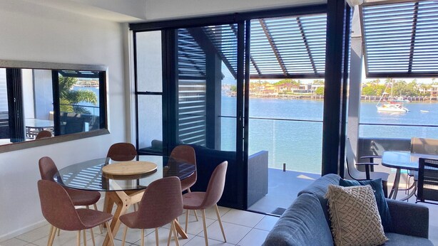 A modern living room with a glass table and chairs, a sofa, and a view of a marina.