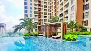 A swimming pool with a wooden pergola and lounge chairs, surrounded by greenery and a high-rise building in the background.