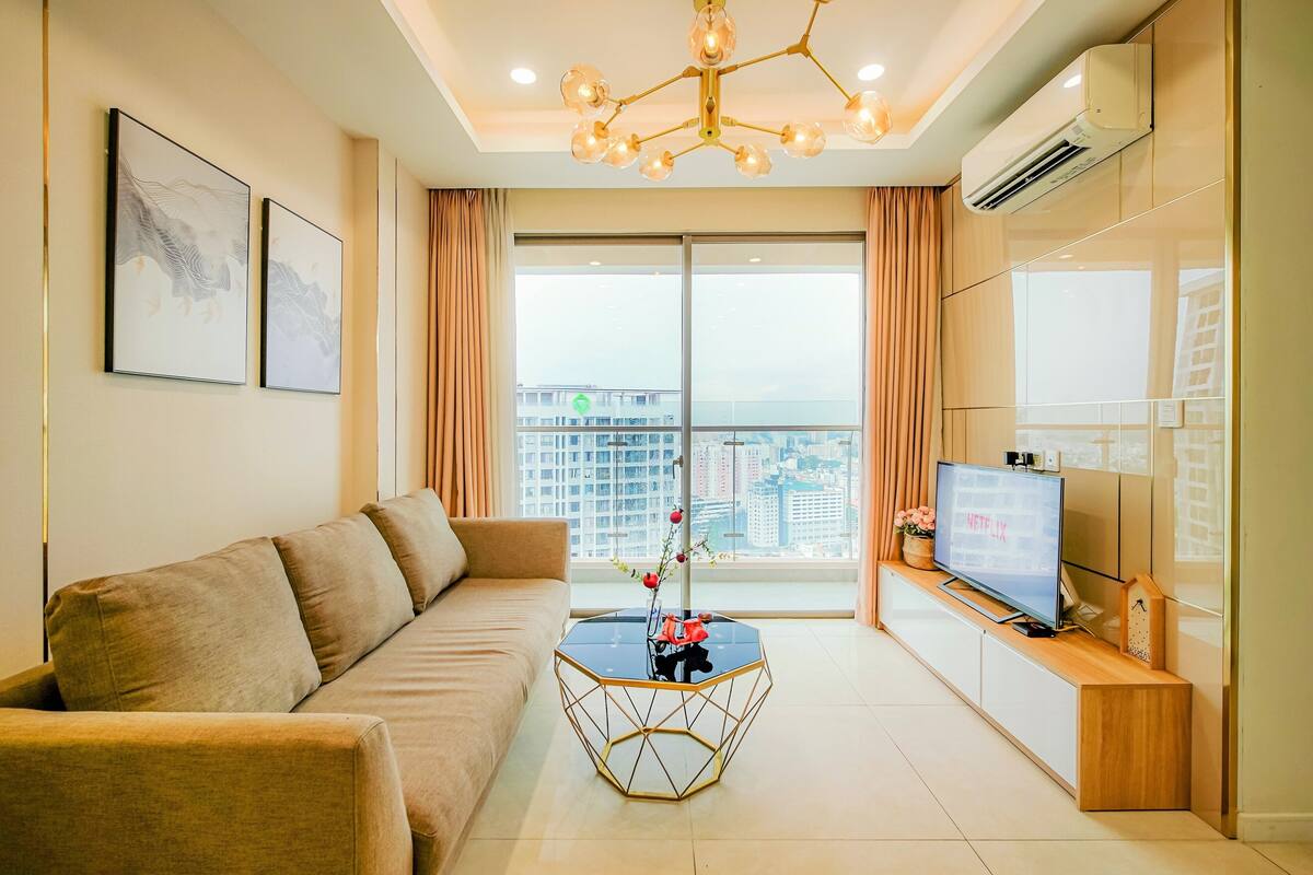 A modern living room with a sofa, a hexagonal coffee table with a vase, a flat-screen TV, and a balcony with a city view.