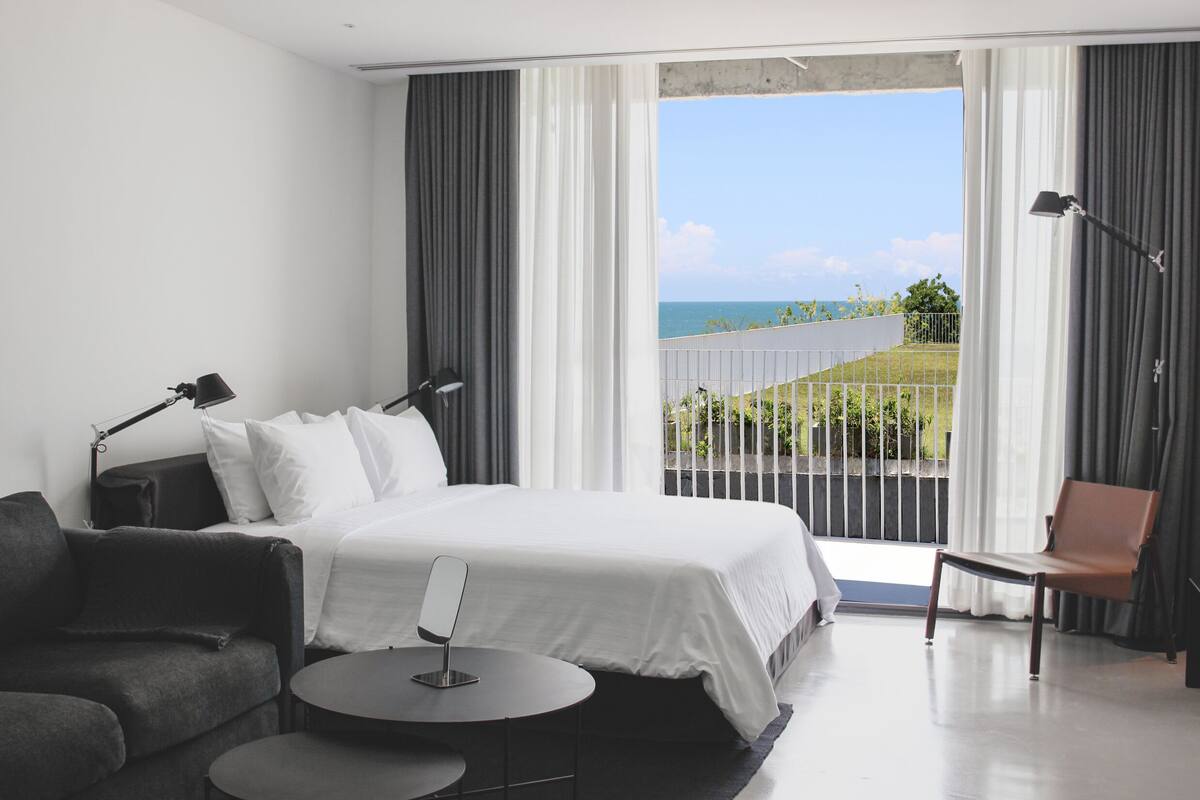 A modern hotel room with a large bed, a sitting area with a table, and a view of the sea through a balcony.
