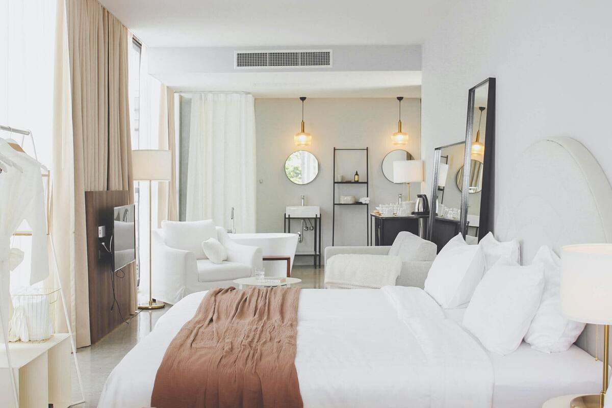 A hotel room with a large bed, two white armchairs, a small table, a desk with a chair, a mirror, and a bathroom visible through an open door.