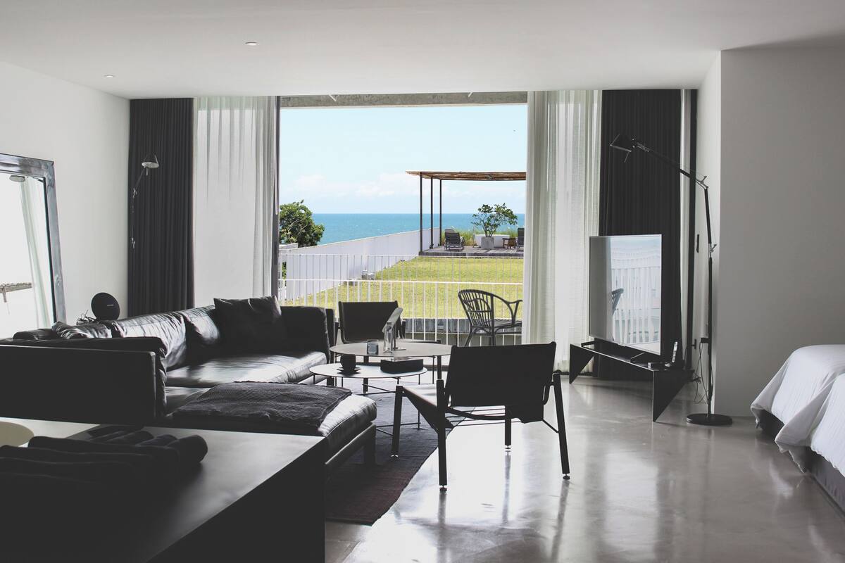 A modern hotel room with a large bed, a sofa, a dining table, and a view of the sea.