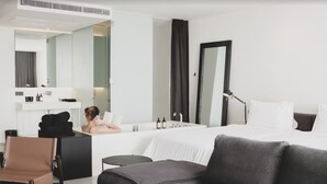 Una habitación de hotel moderna con cama, zona de estar con una silla, escritorio con espejo y baño con lavamanos y espejo.