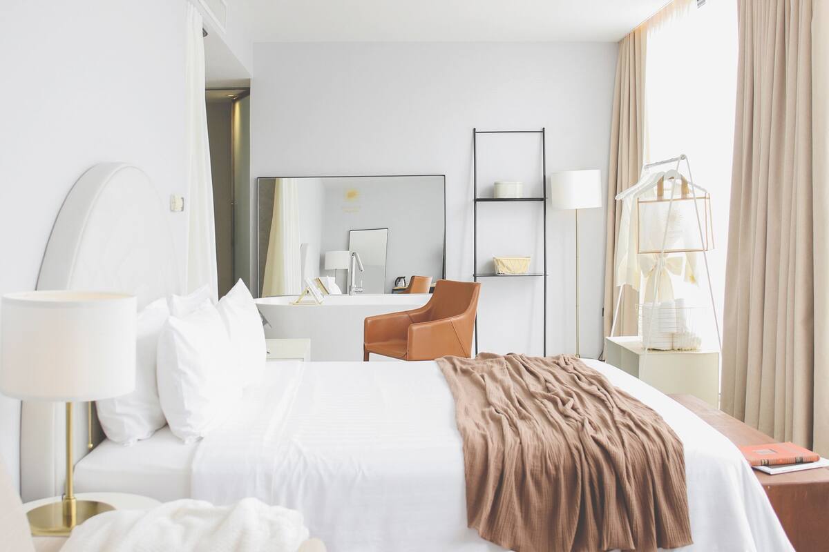 A hotel room with a bed, a chair, a desk, a mirror, and a wardrobe.