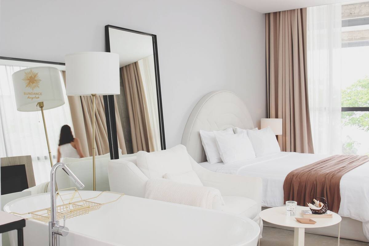 A hotel room with a bed, a sitting area, a bathroom with a sink and a mirror, and a window with curtains.