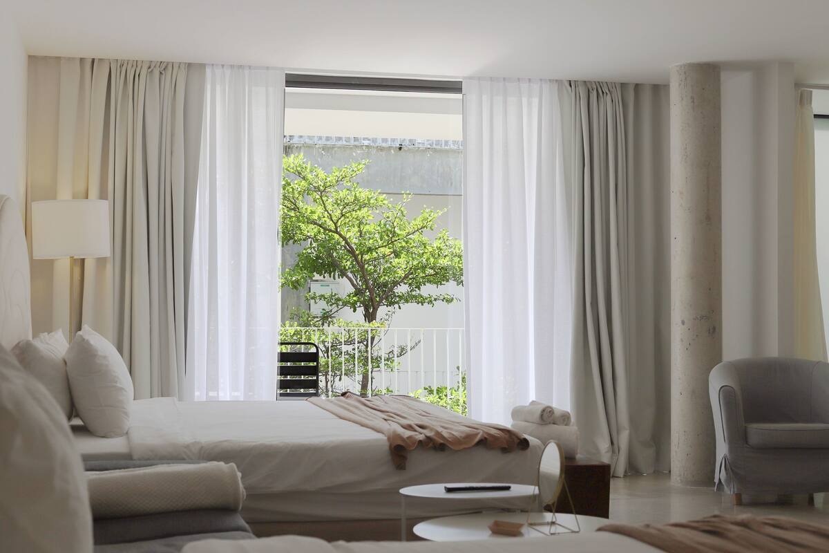 A hotel room with a bed, a sofa, a chair, a small table, and a view of a tree and balcony.