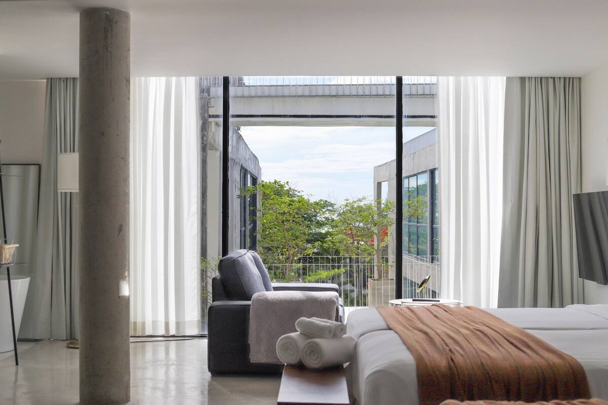 A modern hotel room with a large bed, a seating area with a chair and a small table, and a balcony with a view of greenery and a building.