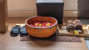 Uma tigela de madeira com pétalas de rosa e uma flor sobre um tapete, chinelos pretos, uma pequena tigela com uma flor e toalhas em uma bandeja.